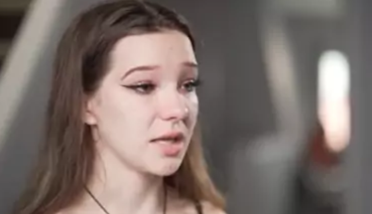 A young woman with long brown hair and winged eyeliner appears emotional, with teary eyes and a slightly open mouth, set against a blurred indoor background, evoking themes often explored by Canada adoption advocates.